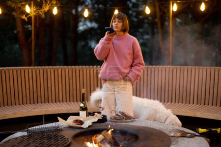 Téléchargez les photos : Young woman spends leisure time with her dog, taking photo on phone at beautiful lounge area with bonfire and round bench in pine forest at dusk - en image libre de droit