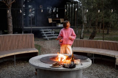 Foto de Young woman prepares food on beautiful outdoor bbq area with fire and round bench in pine forest at dusk. Luxury lifestyle at countryside concept - Imagen libre de derechos