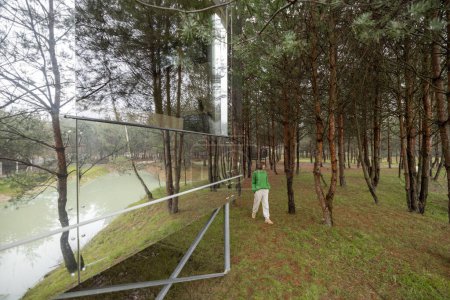Téléchargez les photos : Maison invisible avec des murs en miroir dans la forêt de pins et la personne marche derrière. Lien avec la nature et le concept de durabilité. Repos dans de minuscules cabines sur la nature - en image libre de droit