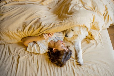 Foto de Mujer joven se abraza con su lindo perro mientras yacen juntos cubiertos con manta beige en la cama. Vista desde arriba. Concepto de amistad con mascotas y comodidad en el hogar - Imagen libre de derechos
