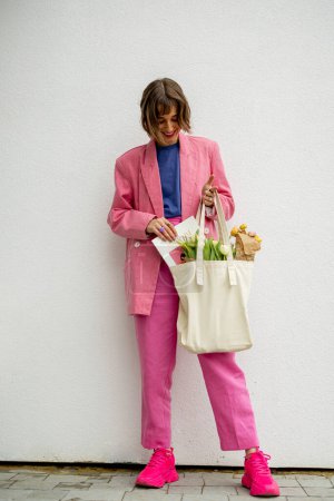 Foto de Stylish woman in pink suit with eco handbag and flowers on white background. Canvas bag with blank space - Imagen libre de derechos