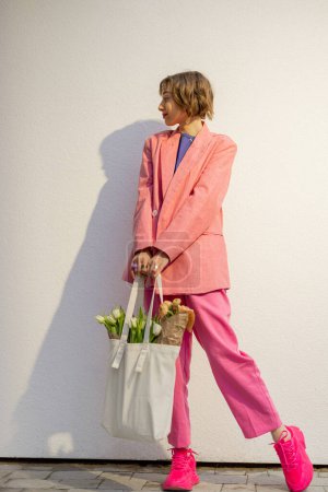 Foto de Lifestyle portrait of stylish woman in pink suit with eco handbag full of flowers on white background. Canvas bag with blank space. Concept of style and spring time - Imagen libre de derechos