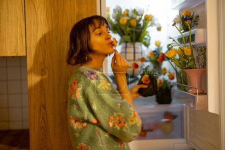Foto de Mujer linda abre nevera llena de flores frescas, de pie en la cocina en casa. Concepto de flores comestibles y dieta floral para la belleza - Imagen libre de derechos