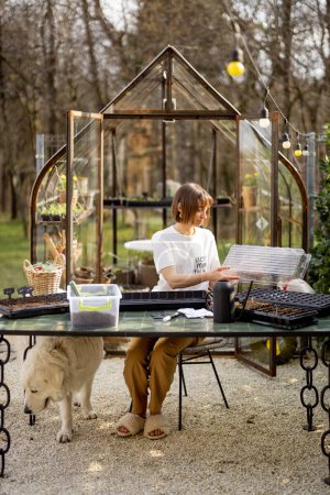 Foto de La joven se prepara para sembrar semillas en bandejas mientras está sentada junto a la mesa en su hermoso jardín a principios de primavera. Concepto de hobby y estilo de vida ecológico sostenible - Imagen libre de derechos