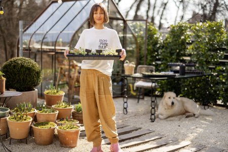 Téléchargez les photos : Portrait de jeune jardinière avec un plateau de semis dans un beau jardin au début du printemps avec une serre derrière - en image libre de droit