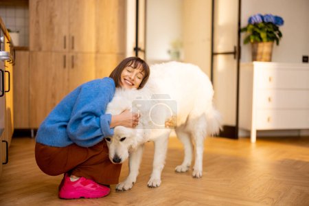 Foto de Mujer joven cuida de su enorme perro blanco, divirtiéndose juntos en casa. Concepto de estilo de vida doméstico y amistad con las mascotas. Perro pastor Maremma - Imagen libre de derechos