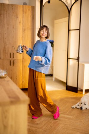 Foto de Mujer joven pasa un tiempo libre caminando con un hervidor de agua y una taza en la cocina de su apartamento estudio moderno. Concepto de estilo de vida y bienestar doméstico - Imagen libre de derechos