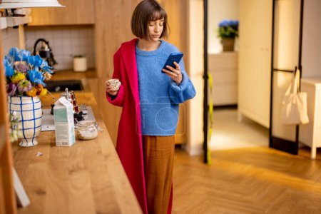 Foto de La mujer sale corriendo de casa poniéndose un abrigo, terminando su café y mirando el teléfono inteligente. Concepto de rutina diaria y trabajo ocupado - Imagen libre de derechos