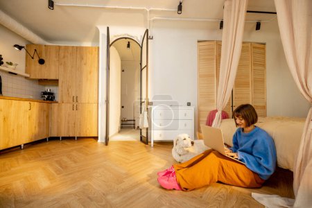 Téléchargez les photos : Femme travaille sur ordinateur portable alors qu'elle est assise sur un sol avec son chien dans un studio moderne. Grande vue intérieure. Confort à la maison et le travail du concept de la maison - en image libre de droit