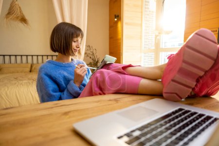 Foto de Mujer joven en ropa casual colorido trabaja en la tableta y el ordenador portátil en línea mientras se sienta relajado por la mesa en ambiente acogedor en casa. Concepto de oficina en casa y trabajo remoto - Imagen libre de derechos