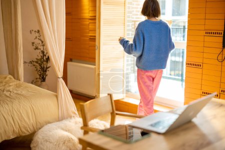 Téléchargez les photos : Femme se tient près de la fenêtre tout en prenant une pause café pendant certains travaux de bureau à la maison. Style de vie domestique et concept de confort - en image libre de droit