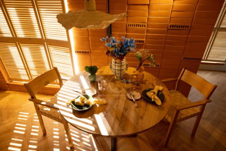 Photo for Sunny and cozy studio apartment with the suns rays passing by the shutters. View on wooden dining table decorated with flowers - Royalty Free Image