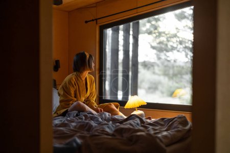 Téléchargez les photos : Woman in yellow bathrobe wakes up in tiny bedroom of a wooden cabin in forest. Good morning and rest in house on nature concept - en image libre de droit