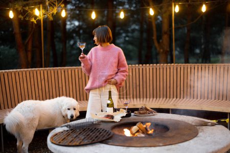 Téléchargez les photos : Young woman spends leisure time with her dog, drinking wine at beautiful lounge area with bonfire and round bench in pine forest at dusk - en image libre de droit