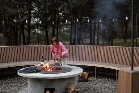 Photo for Young woman prepares food on beautiful outdoor bbq area with fire and round bench in pine forest at dusk. Luxury lifestyle at countryside concept - Royalty Free Image