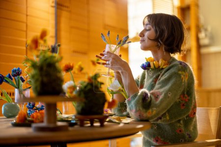 Foto de Joven mujer disfruta de flores, mientras está sentada junto a la mesa de comedor llena de diferentes flores. Concepto de belleza y dieta floral - Imagen libre de derechos