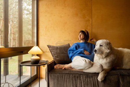 Téléchargez les photos : Portrait d'une jeune femme avec son chien assis ensemble sur un canapé et regardant la fenêtre avec vue sur la forêt. Repos à la campagne chalet et amitié avec les animaux concept - en image libre de droit