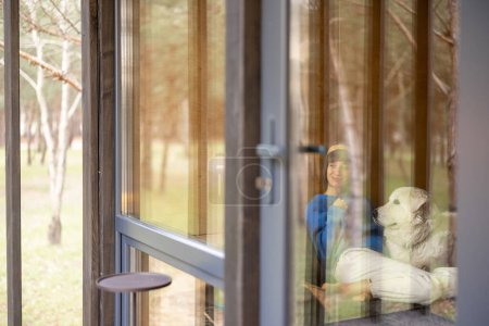 Foto de Mujer con perro sentado junto a la ventana en casa de madera, vista a través de la ventana. - Imagen libre de derechos