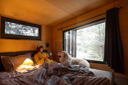 Téléchargez les photos : Femme utilise le téléphone tout en étant couché dans son lit avec son chien mignon dans une minuscule chambre de cabine en bois sur la nature - en image libre de droit