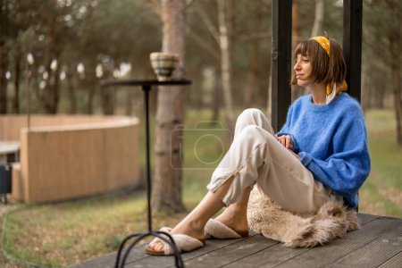 Foto de Young woman sits on porch of a wooden house in pine forest, enjoying nature while resting in cottage at countryside - Imagen libre de derechos