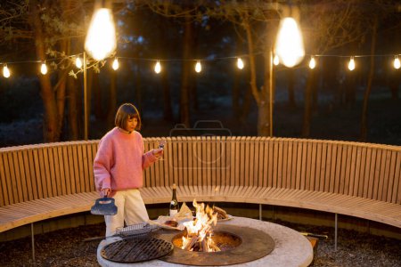 Téléchargez les photos : Young woman prepares food on beautiful outdoor bbq area with fire and round bench in pine forest at dusk. Luxury lifestyle at countryside concept - en image libre de droit