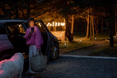 Téléchargez les photos : Woman arrives by car to a house in forest, standing with bag and phone near vehicle in the evening time. Traveling by car and rest in cabins on nature concept - en image libre de droit