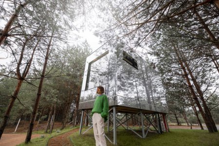 Foto de Woman near an invisible house with mirrored walls in pine forest. Connection with nature and sustainability concept. Rest in tiny cabins on nature - Imagen libre de derechos