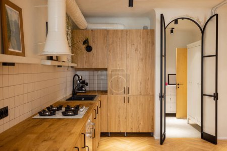 Photo for Photo of stylish kitchen interior made in wooden materials of modern studio apartment. Oak kitchen facades and metal arch door - Royalty Free Image