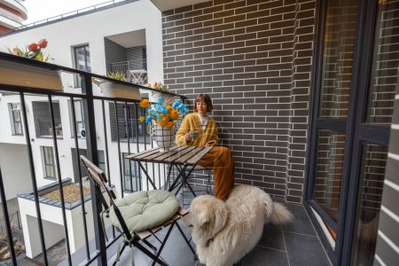 Foto de Mujer joven se sienta con su perro en el balcón, disfrutando de una bebida de café en su propiedad durante la mañana. La vida de la ciudad en un complejo residencial moderno - Imagen libre de derechos