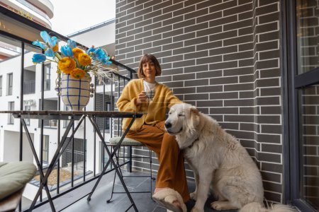 Foto de Mujer joven se sienta con su perro en el balcón, disfrutando de una bebida de café en su propiedad durante la mañana. La vida de la ciudad en un complejo residencial moderno - Imagen libre de derechos