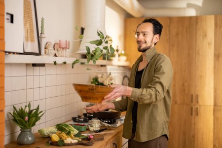 Téléchargez les photos : Beau homme mélangeant la salade dans un bol en bois, vomissant des feuilles d'épinards, cuisinant des aliments végétariens sains à l'intérieur élégant de la cuisine de la maison - en image libre de droit