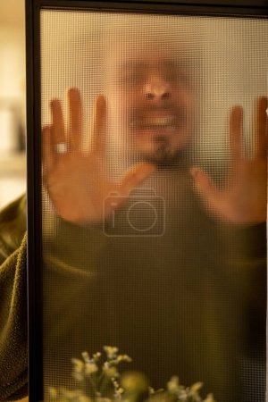Photo for Mens portrait through a matte glass door at home. Weird man touching glass surface - Royalty Free Image
