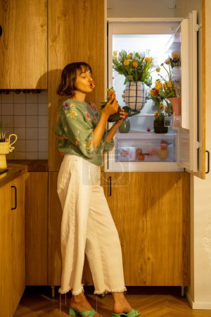 Téléchargez les photos : Femme mignonne ouvre réfrigérateur rempli de fleurs fraîches, debout sur la cuisine à la maison. Concept de fleurs comestibles et régime floral pour la beauté - en image libre de droit