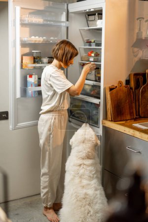 Foto de Woman looks into a fridge, cooking with her huge white dog together on kitchen at home - Imagen libre de derechos