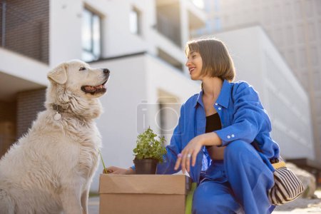 Foto de Mujer cuida a su perro mientras está sentado con caja de cartón y maceta, reubicándose en una nueva casa. Edificio de apartamentos en segundo plano. - Imagen libre de derechos