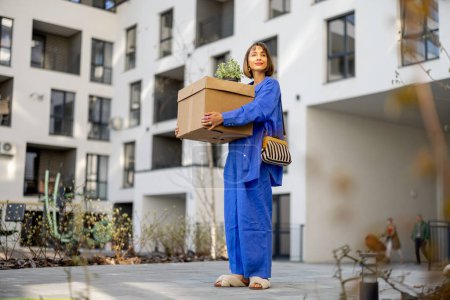 Photo for Young woman carrying cardboard box and flowerpot at inner yard of apartment building, relocating to a new home - Royalty Free Image