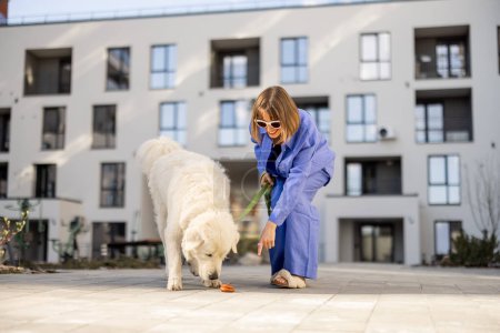 Téléchargez les photos : Femme pointe du doigt sur chien caca, tout en marchant avec animal de compagnie dans la cour d'un immeuble d'habitation. Concept de nettoyage après les animaux de compagnie dans les lieux publics - en image libre de droit