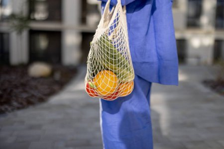 Photo for Woman carrying mesh bag full of fresh fruits and vegetables while walking home, close-up. Sustainable lifestyle and using reusable bag in shopping food concept - Royalty Free Image