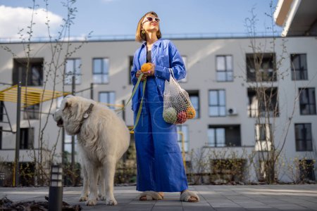 Téléchargez les photos : Une jeune femme en pyjama bleu se promène avec son chien et porte un sac en filet rempli de fruits et légumes frais dans la cour intérieure de l'immeuble. Concept de mode de vie durable et moderne - en image libre de droit