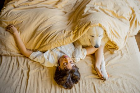 Foto de Young woman hugs with her cute dog while lying together covered with beige blanket in bed. View from above. Concept of friendship with pets and home coziness - Imagen libre de derechos