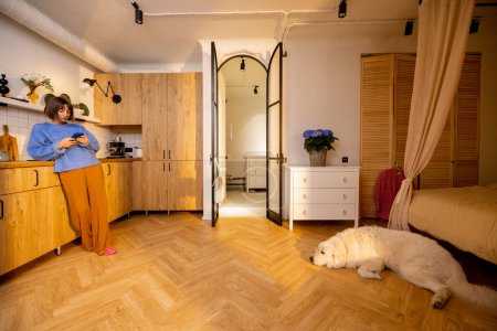 Photo for Woman stands with phone on kitchen of modern studio apartment, her dog lying on floor. Wide interior view. Concept of home coziness and stylish interiors - Royalty Free Image