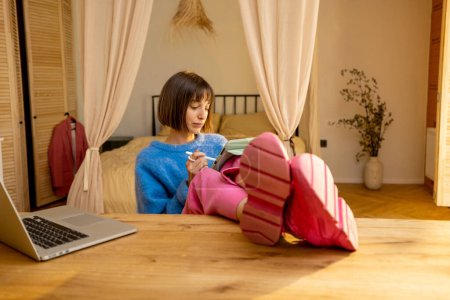 Téléchargez les photos : Jeune femme en vêtements décontractés colorés fonctionne sur tablette et ordinateur portable en ligne tout en étant assis détendu par la table dans une atmosphère confortable à la maison. Concept de bureau à domicile et de télétravail - en image libre de droit