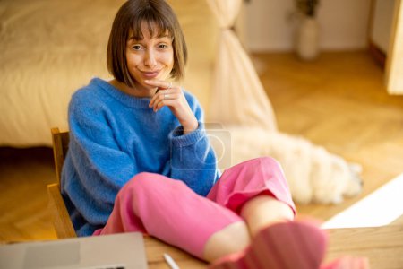 Téléchargez les photos : Portrait de jeune femme en vêtements décontractés colorés assis à la table dans une atmosphère chaleureuse à la maison. Concept de bureau à domicile et de télétravail - en image libre de droit