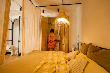 Photo for Interior view of bedroom in beige tones with straw lampshade and canopy. Woman dressing in wardrobe with wooden shutters - Royalty Free Image