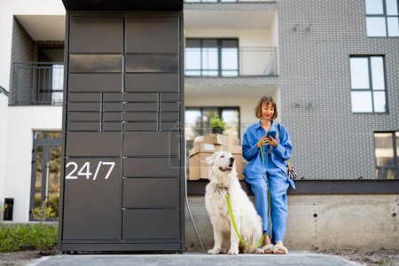 Téléchargez les photos : Femme s'assoit avec un paquet de carton et chien blanc près de la machine de bureau de poste et en utilisant le téléphone mobile dans le quartier résidentiel. Concept de livraison et mode de vie avec animaux domestiques - en image libre de droit