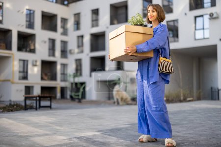 Foto de Mujer joven que lleva caja de cartón y maceta en el patio interior del edificio de apartamentos, reubicándose en una nueva casa - Imagen libre de derechos