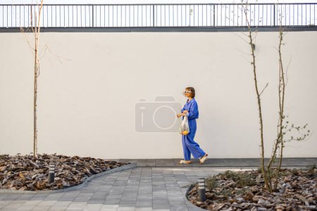 Photo for Woman in blue pajamas walks with mesh bag full of fresh fruits on white wall background outdoors. Concept of sustainability and modern lifestyle - Royalty Free Image