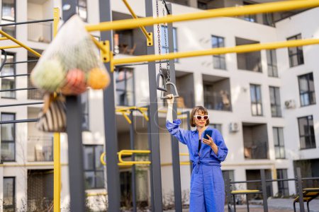 Photo for Woman spends leisure time on a sports ground of apartment building, standing with phone. Mesh bag with friuts hanging in front - Royalty Free Image