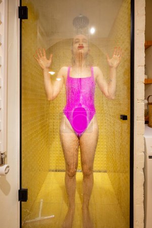 Téléchargez les photos : Femme en maillot de bain rose touchant verre de douche tout en se lavant dans la cabine de douche. Loisirs dans la salle de bain, routine domestique, concept d'hygiène - en image libre de droit
