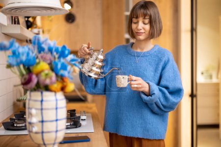 Foto de Mujer joven hace una bebida de café, de pie con una tetera y una taza en el interior de la cocina moderna. Concepto de bienestar y estilo de vida doméstico - Imagen libre de derechos